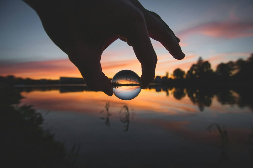 Une bulle d'eau tenue entre le pouce et l'index d'une main, devant un paysage avec un coucher de soleil