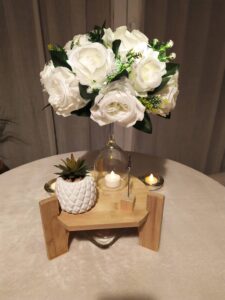 Centre de table pour un dîner romantique, composé d'un joli support en bois, d'un bouquet de roses blanches, de bougies et d'une mini plante verte