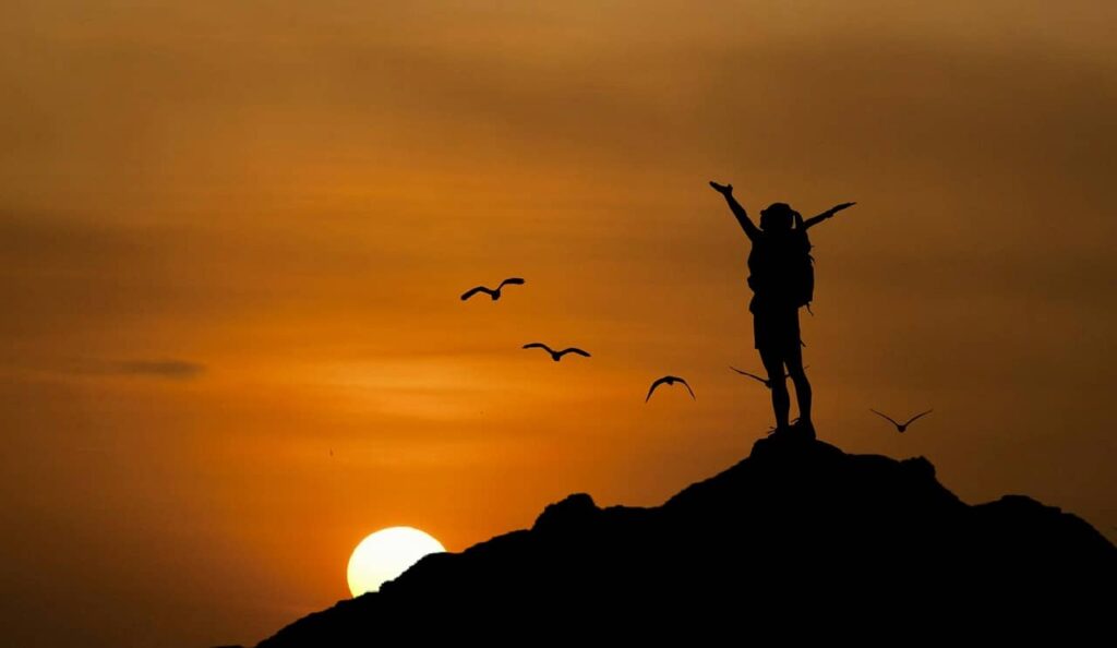 Jeune fille levant les bras au ciel, en haut d'une montagne et devant un coucher de soleil