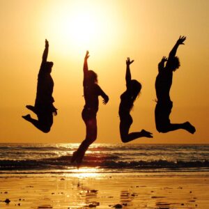 Groupe de 4 amis, sautant de joie sur une plage, devant un coucher de soleil